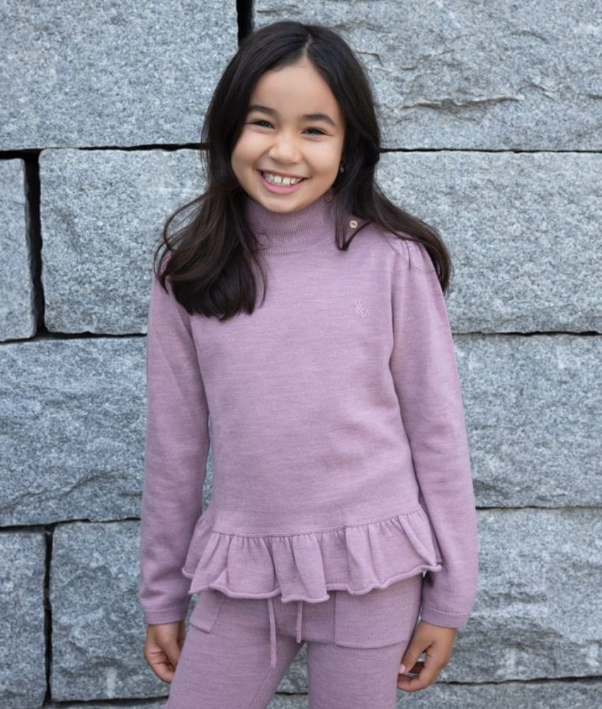 Rosa strikket ullgenser fra Vilje & Ve med høy hals og knappedetaljer, komplettert med en sjarmerende rysjekant nederst. Perfekt for en varm og stilig look og er superfin til Aspen strikket bukse.